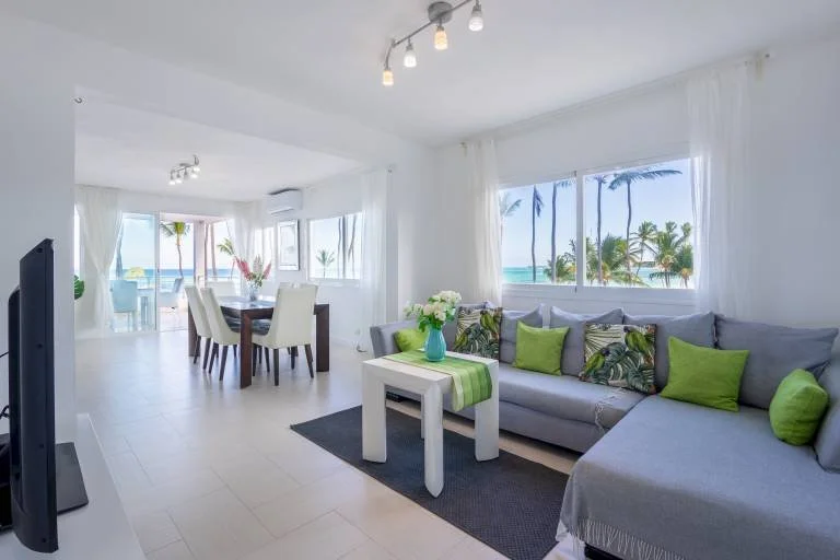 Salon moderne avec canapé gris, coussins verts, table avec vase et vue sur la mer par les fenêtres.