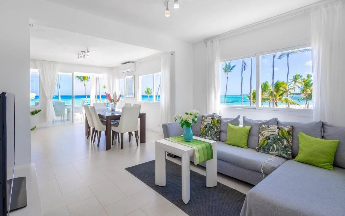 Salon moderne avec décoration tropicale, canapé gris, coussins verts, table basse blanche, grande table à manger avec chaises blanches, grandes fenêtres offrant vue sur la plage et les palmiers.