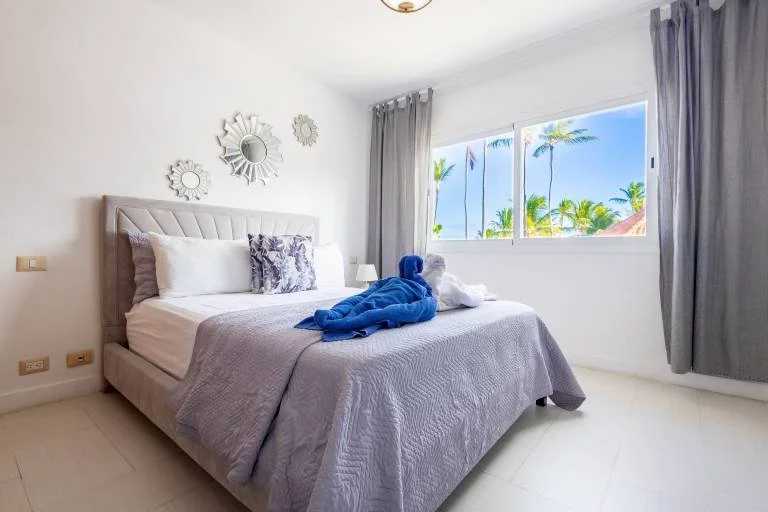 Chambre lumineuse avec un lit double décoré de coussins et d'une serviette de bain en forme de cygne bleu. Fenêtre montrant des palmiers à l'extérieur. Mur orné de miroirs décoratifs.