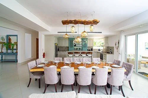 Salle à manger moderne avec grande table bois et chaises rembourrées grises, cuisine ouverte en arrière-plan et lustre en bois avec ampoules suspendues.