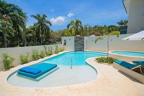 Piscine extérieure avec transats, coussins bleus, palmiers, et ciel ensoleillé.