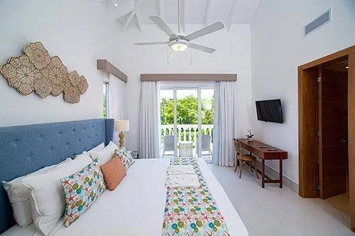 Chambre lumineuse avec lit double, tête de lit bleue, coussins colorés, ventilateur au plafond, télévision murale, bureau en bois et porte-fenêtre donnant sur un balcon.