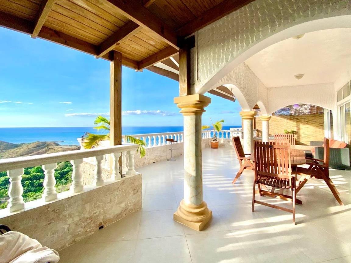 Terrasse avec arches surplombant une vue panoramique sur la mer, meubles en bois, et soutien de colonnes décoratives.