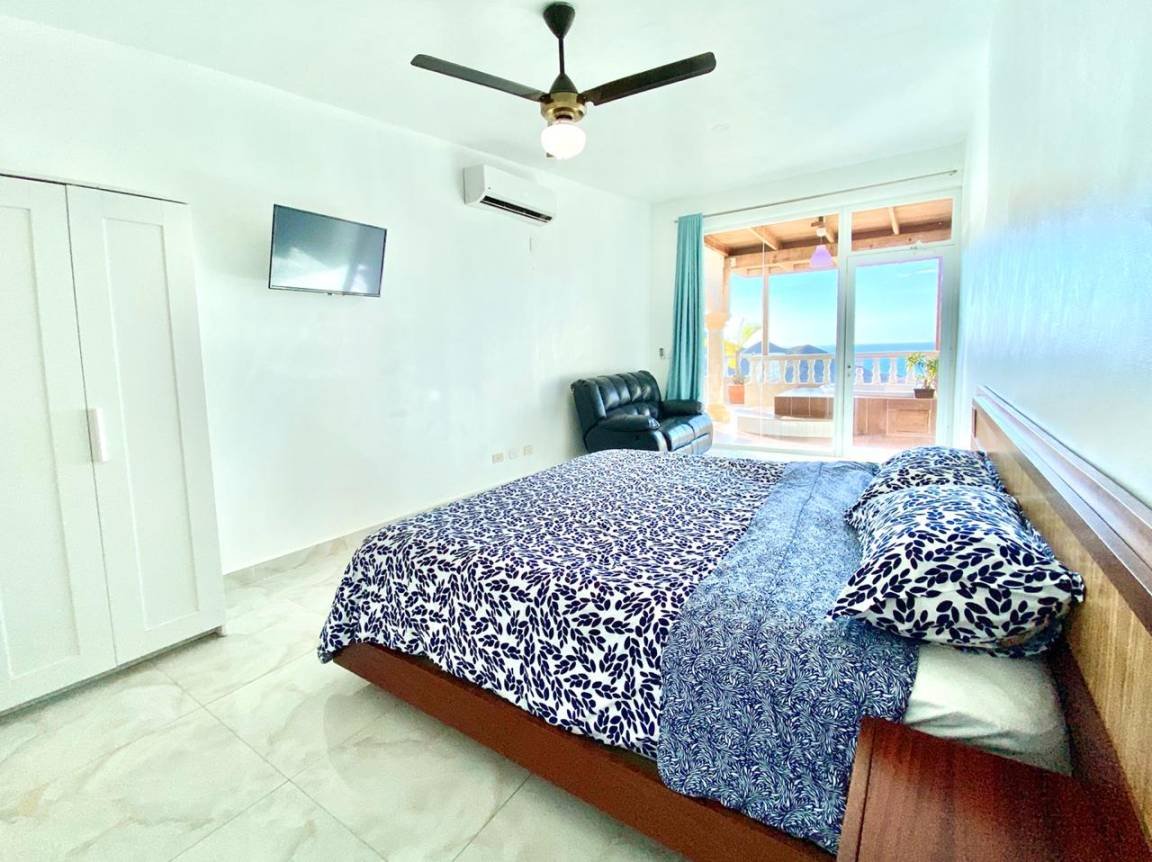 Chambre avec lit double, couverture à motif bleu, téléviseur mural, fauteuil noir, ventilateur de plafond, climatisation, et portes-fenêtres menant à un balcon avec vue sur la mer."}