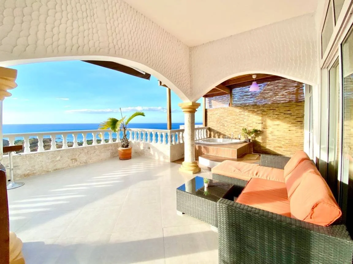 Terrasse couverte avec vue sur la mer, mobilier de jardin, canapé avec coussins orange, pot de plante, jacuzzi et piliers décoratifs. Atmosphère lumineuse et ensoleillée.