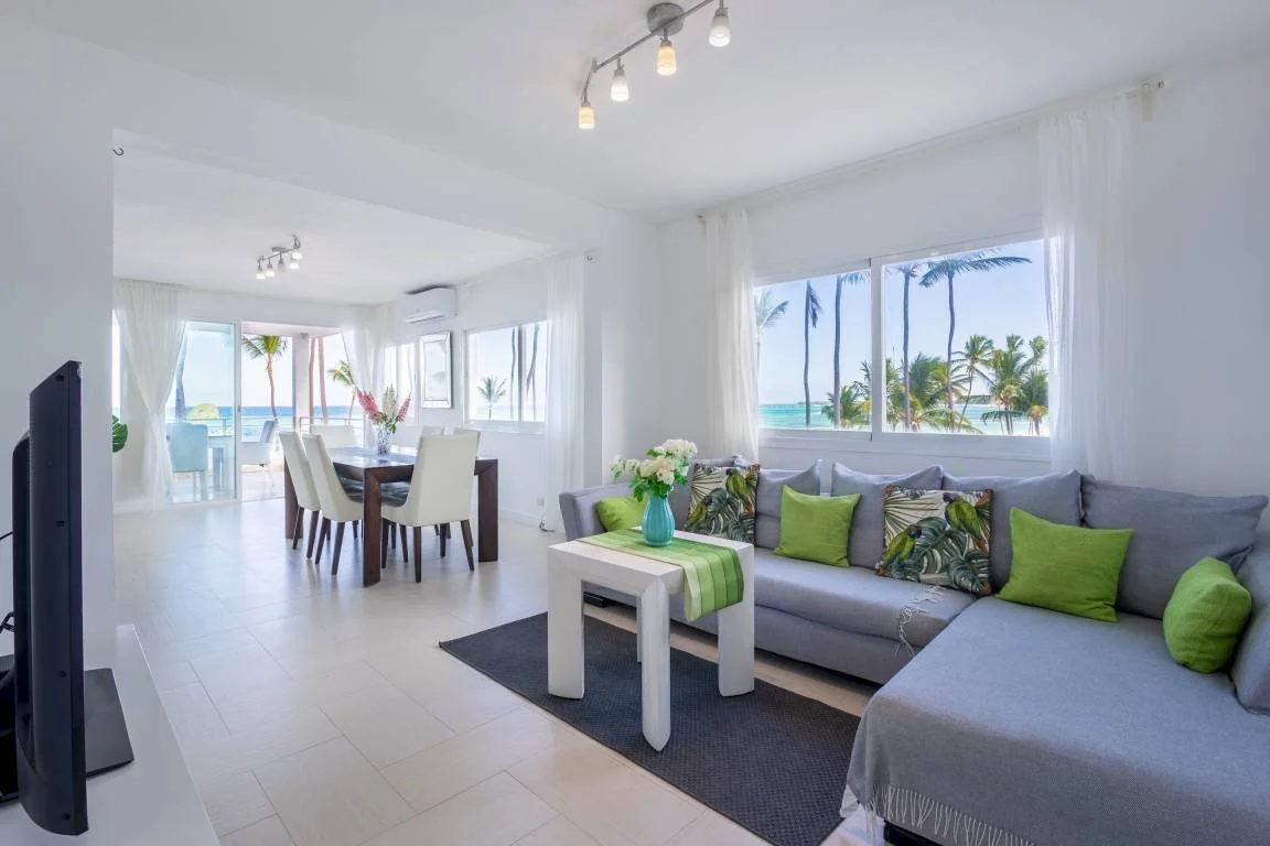 Intérieur moderne et lumineux avec un salon équipé de canapés gris, coussins verts et décorations florales, ainsi qu'une salle à manger avec vue sur la mer et des palmiers par les grandes fenêtres.