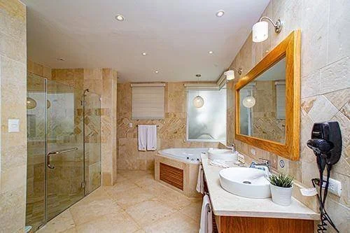 Salle de bain moderne avec douche en verre, baignoire, vasque et miroir, murs en carrelage beige, sèche-cheveux mural et serviettes blanches.