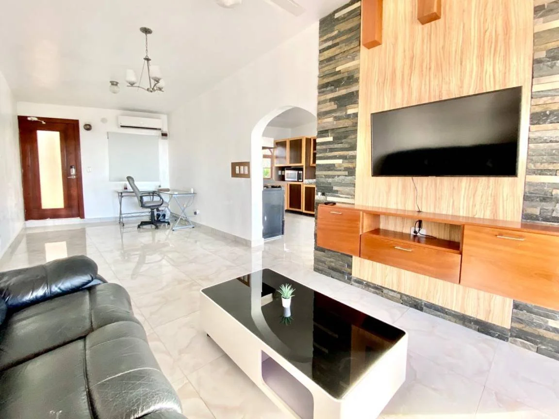 Salon moderne avec canapé en cuir noir, télévision murale, table basse, et bureau avec chaise. Décor contemporain avec murs en pierre et bois.