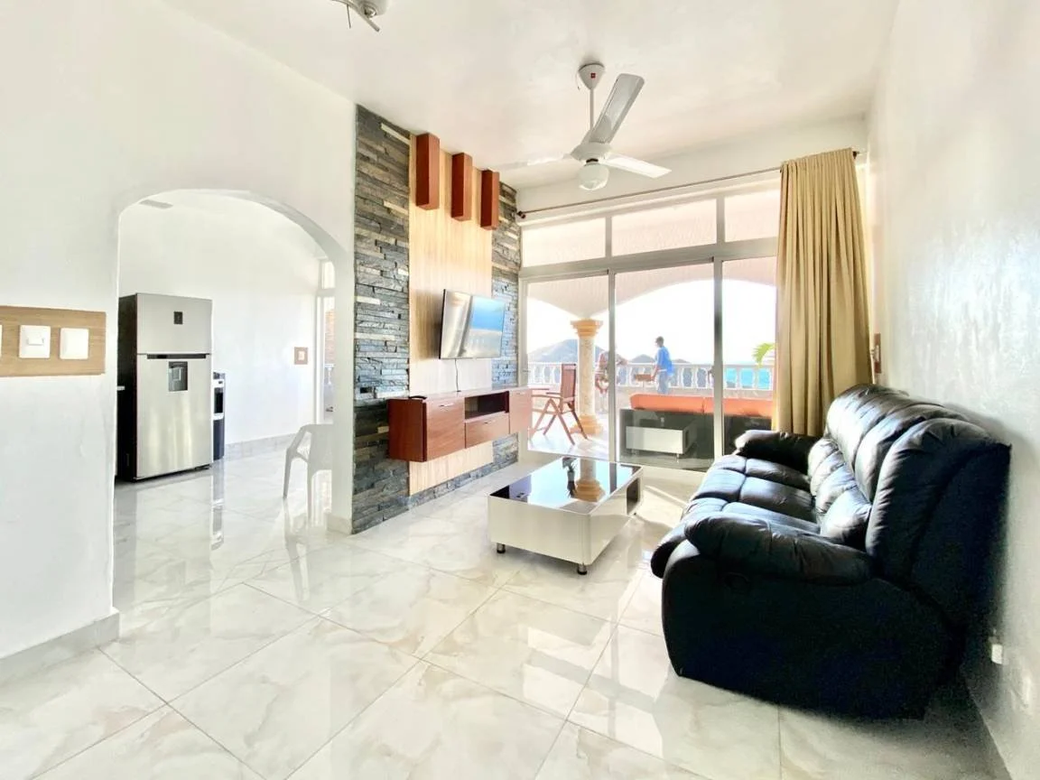 Salon moderne avec canapé en cuir noir, table basse en verre, télévision murale, ventilateur de plafond, grande fenêtre avec rideaux, et cuisine avec réfrigérateur visible.