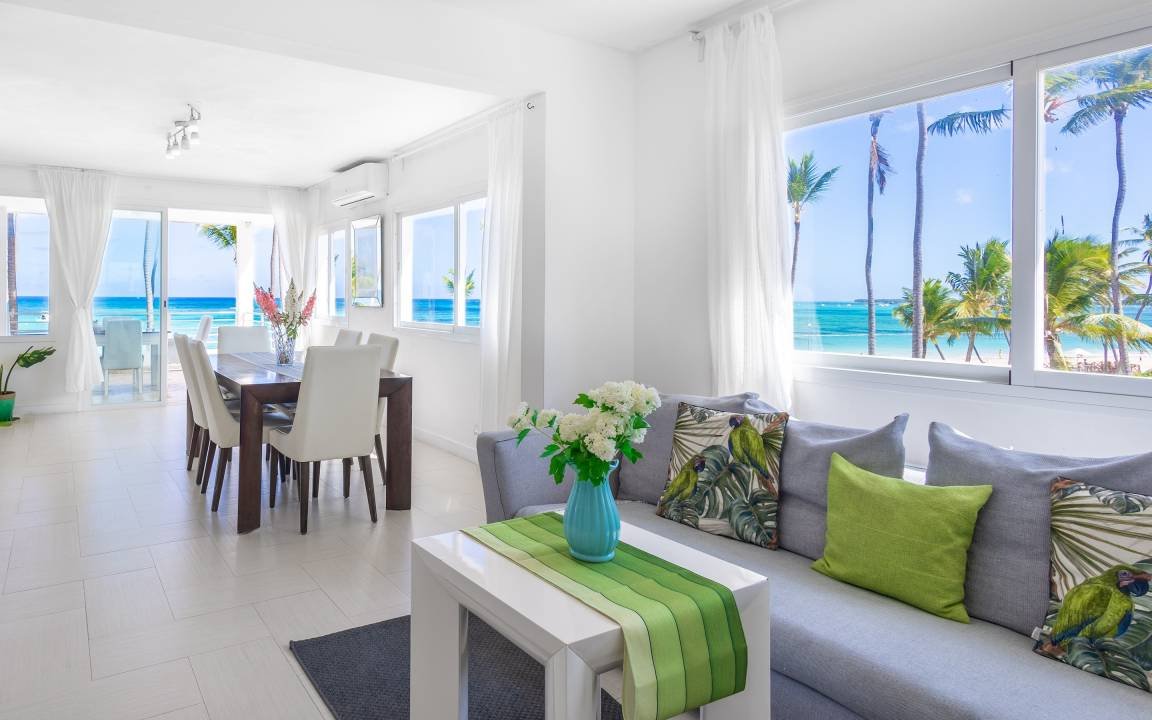 Intérieur lumineux d'une maison en bord de mer avec vue sur l'océan. Mobilier moderne, canapés gris, coussins verts à motifs, table à manger en bois avec chaises blanches, et décoration florale. Grandes fenêtres avec rideaux blancs offrant une vue su