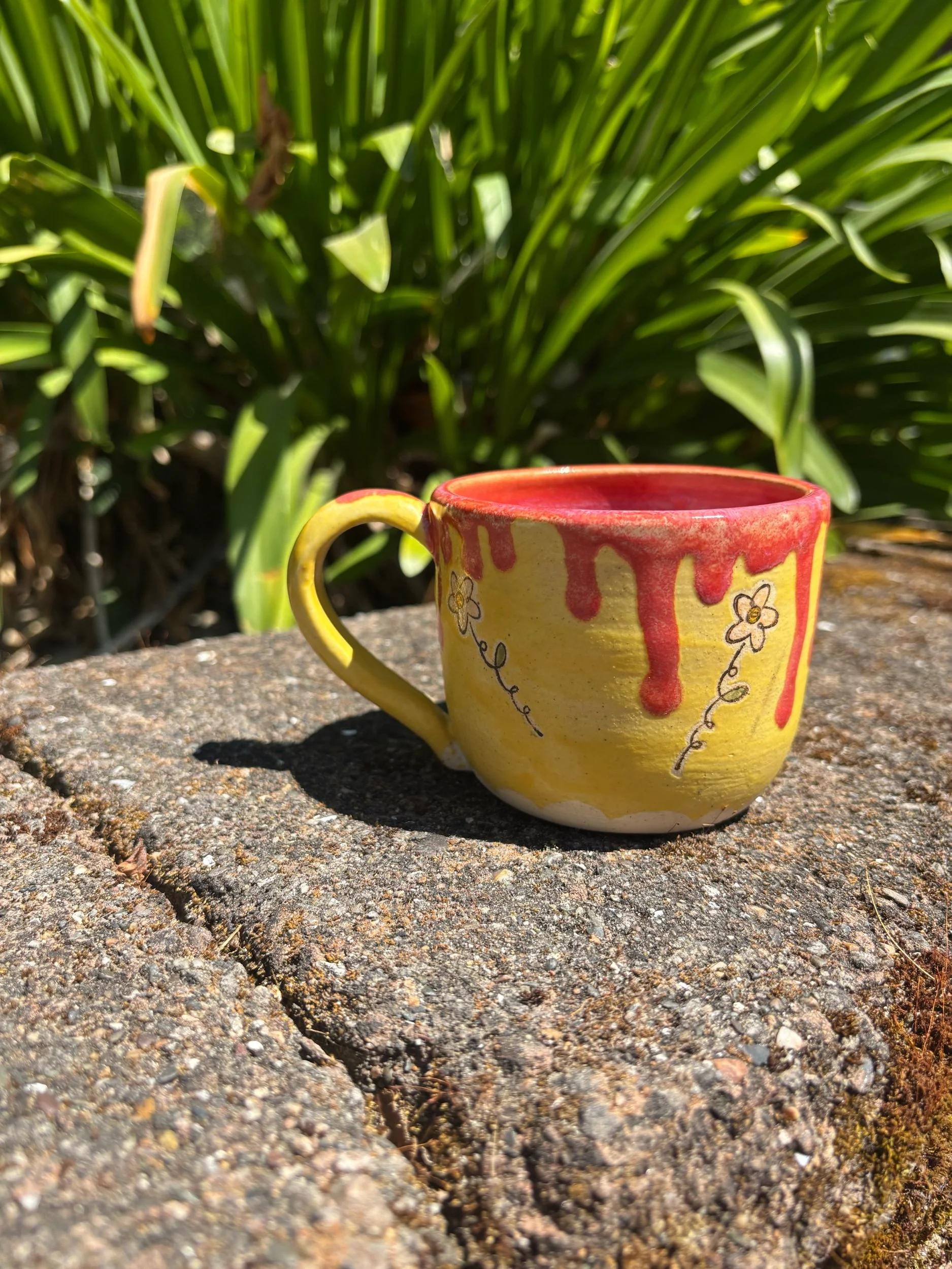 A yellow ceramic mug with red dripping glaze on top, decorated with small hand-painted flowers and vines, sitting on a rough stone surface outdoors with green foliage in the background.