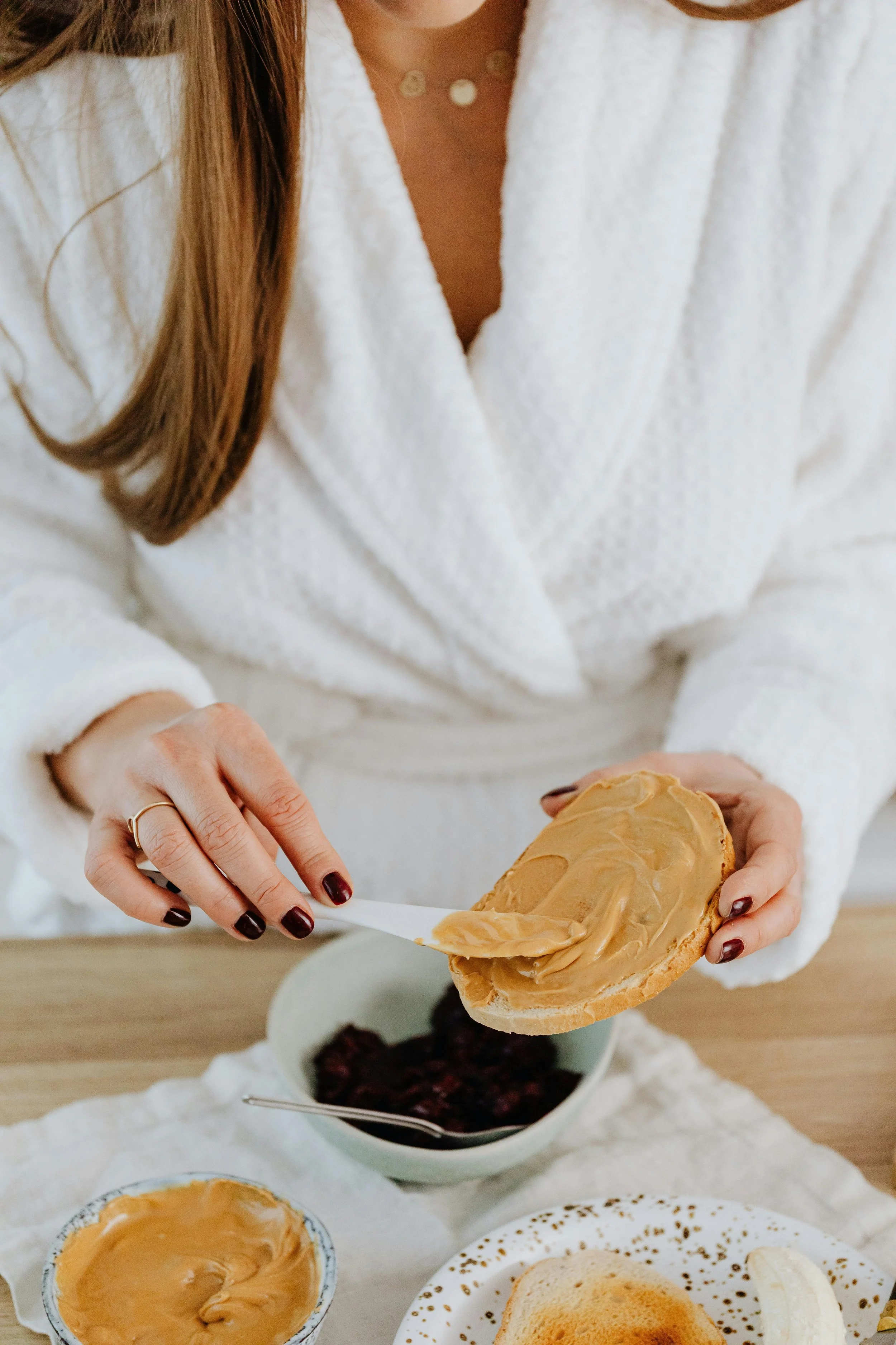 Person in a white robe spreading peanut butter on toast with a knife, with a bowl of jam nearby.