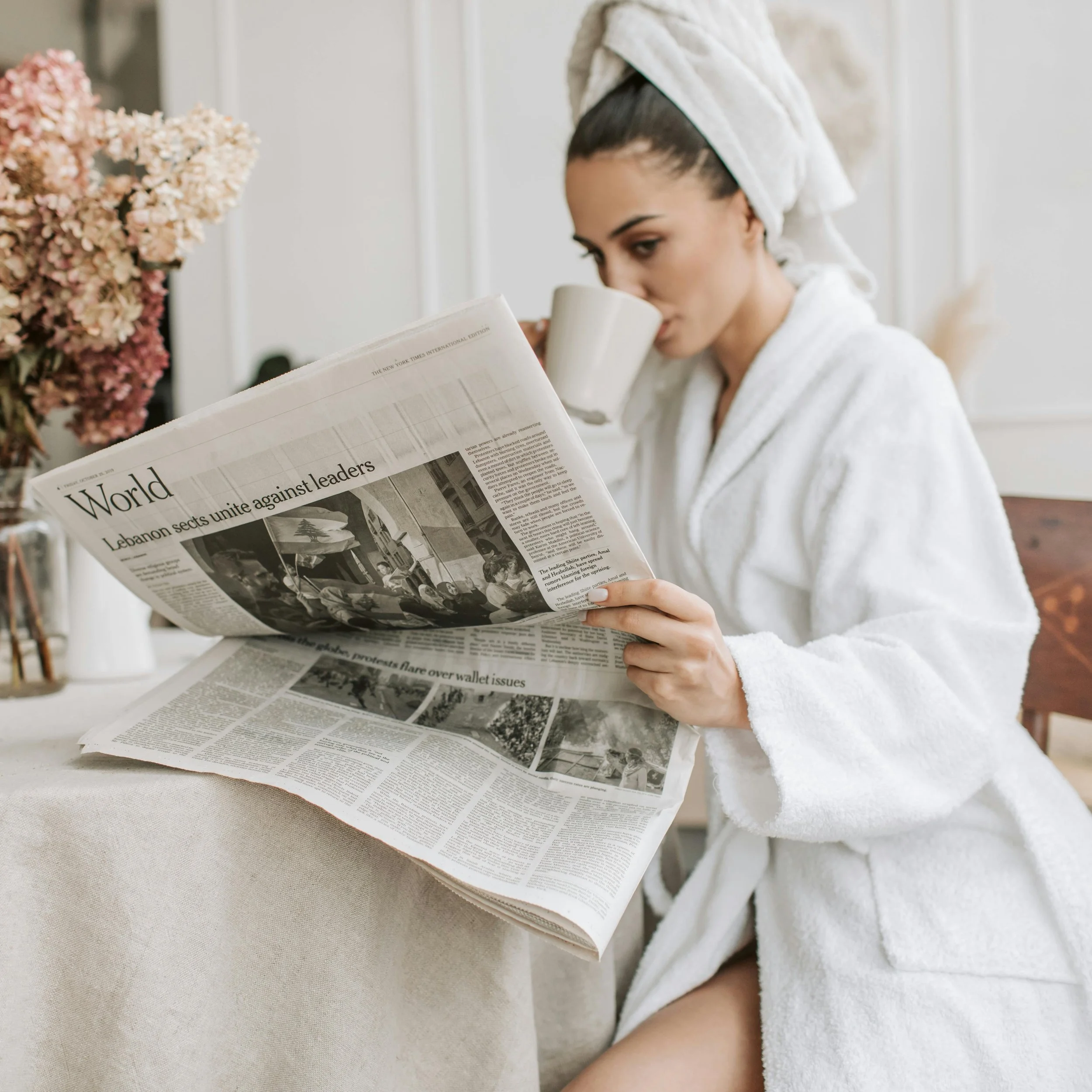 Person in a white robe with a towel on their head, drinking from a mug and reading a newspaper, sitting near a table with flowers.