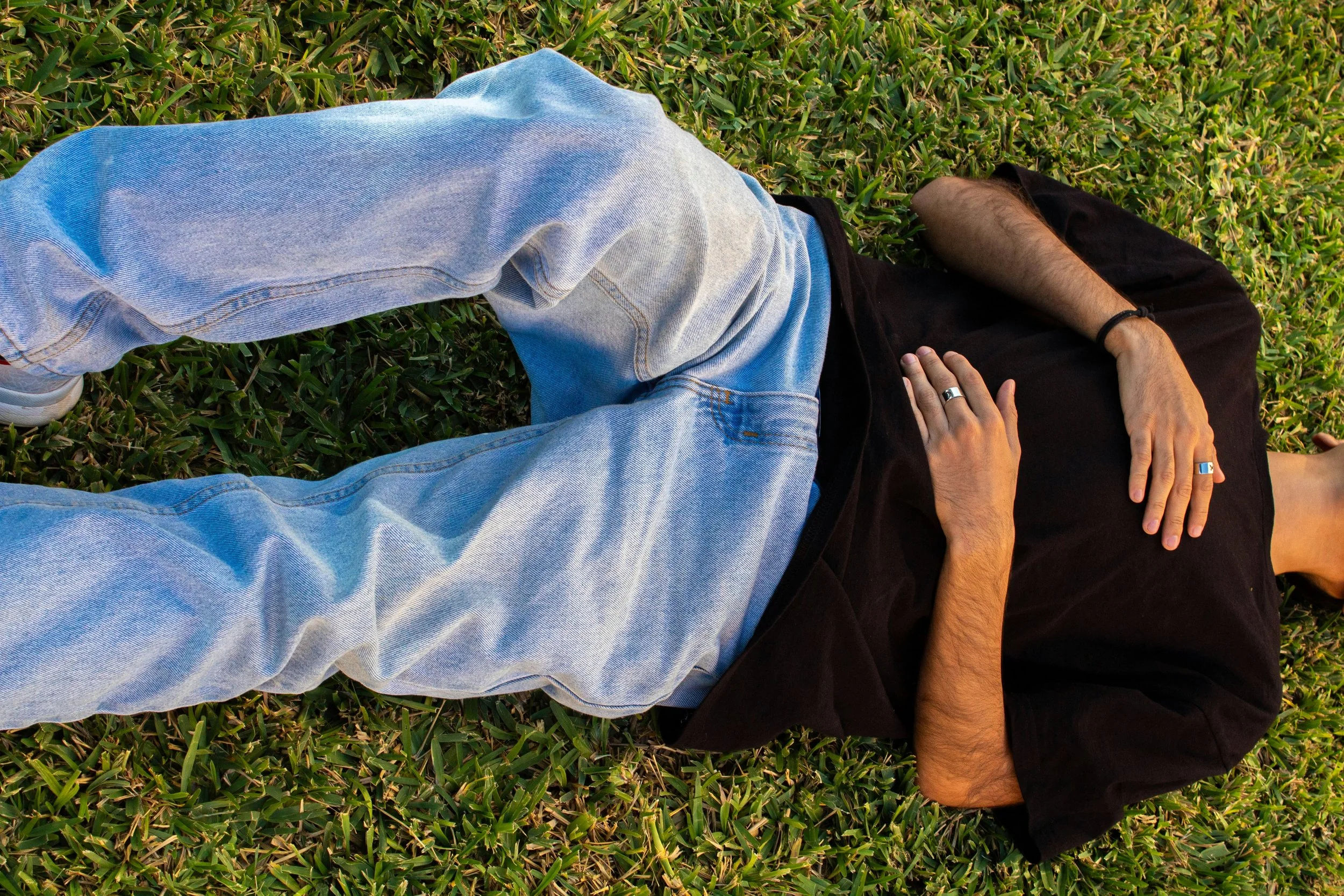 Person lying on the grass wearing a black t-shirt and jeans with their hands on their belly and chest