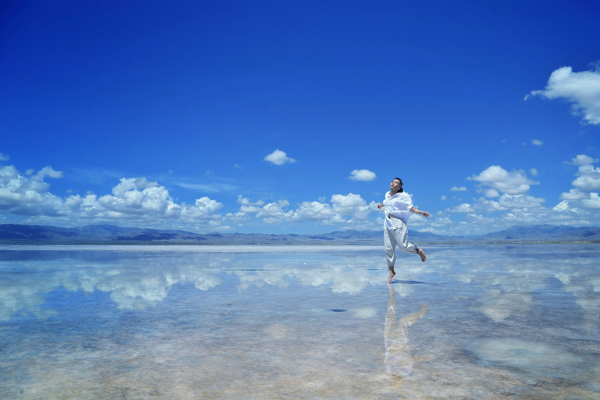 person of chinese decent dressed in all white running on the beach arms open blue sky with puffy clouds and mountains in the background water in the foreground with reflections of the clouds