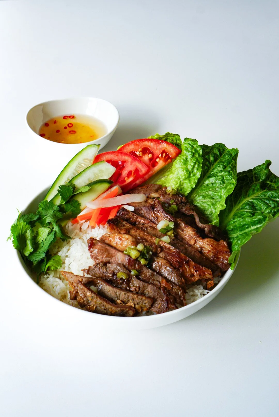 Warm rice bowl topped with grilled beef, topped with fresh veggies, and a side of fish sauce