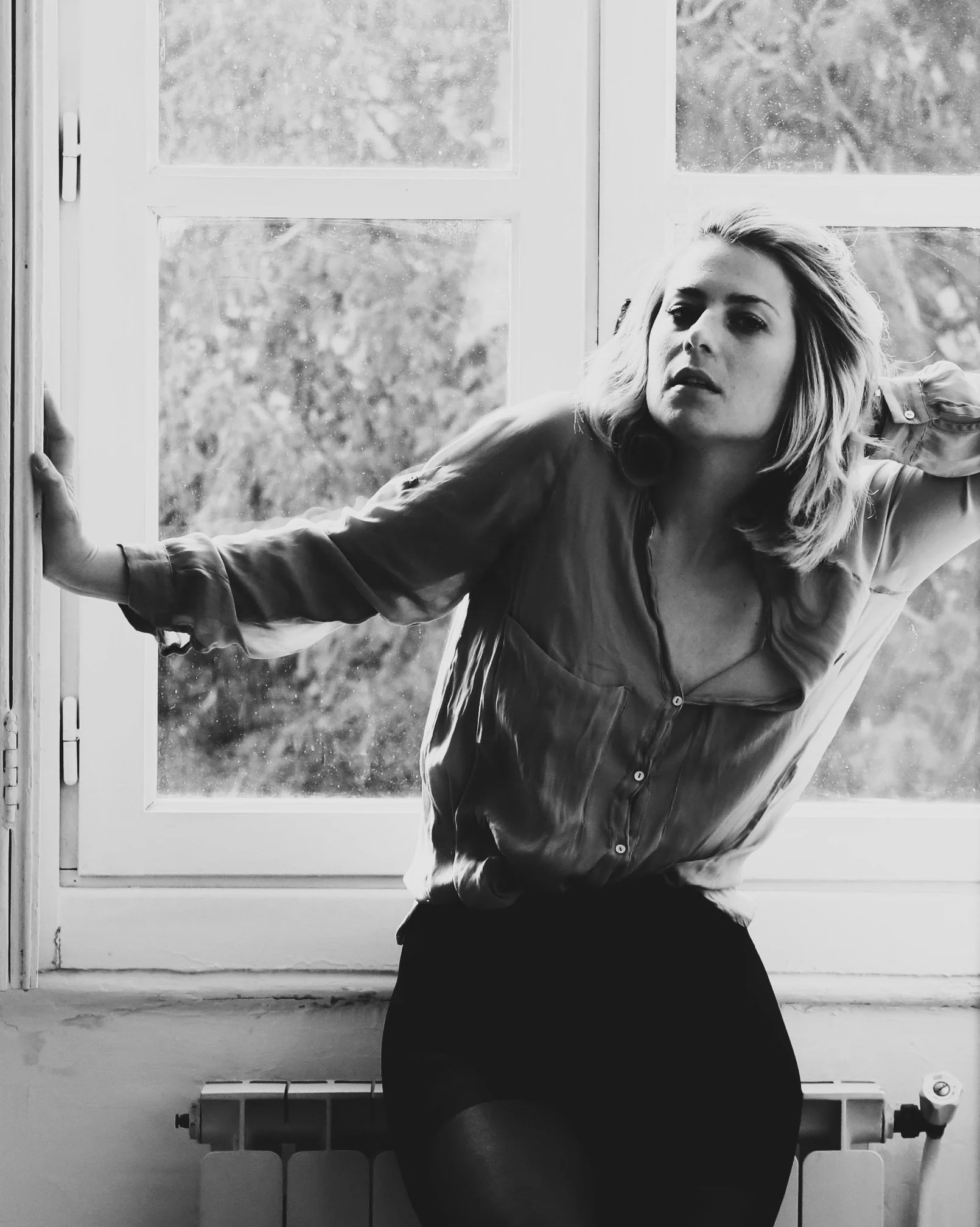 Black and white photo of a woman sitting on a window sill, looking away from the camera, with her hand resting on the window frame and the other behind her head.