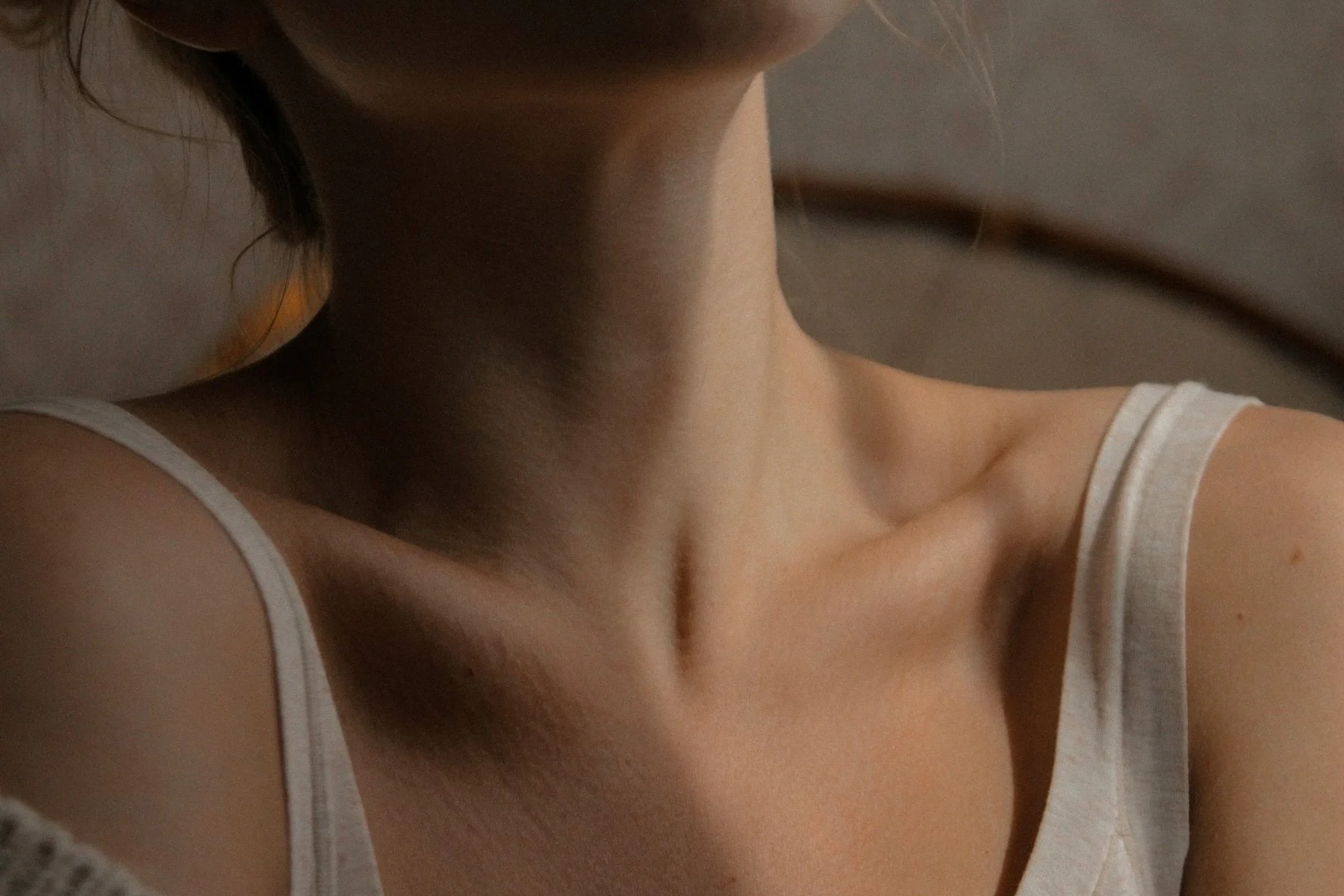 Close-up photo of a woman's neck, collarbone, and shoulder in a white tank top, with a part of her face and hair visible.