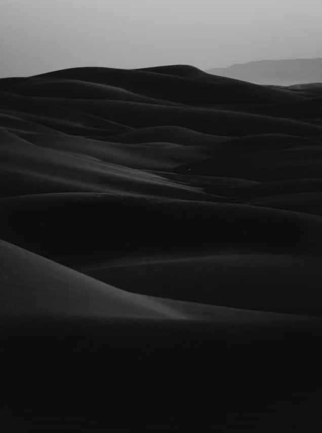 Black and white photo of sand dunes with smooth, curved shapes under a cloudy sky.