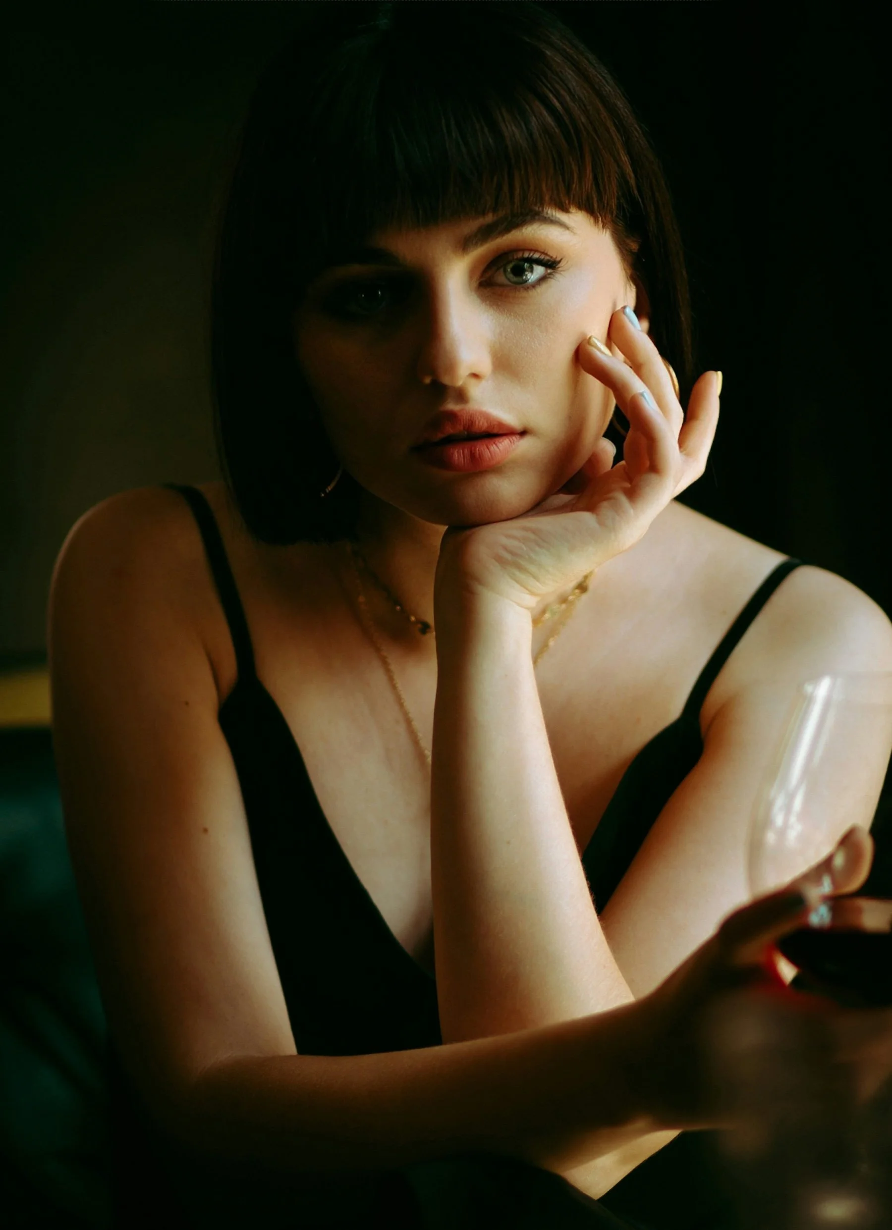 A young woman with short dark hair and bangs, wearing a black spaghetti strap top, is resting her chin on her hand while looking at the camera. She has blue eyes and neutral makeup, with subtle jewelry. The background is dark, with a soft light illuminating her face.