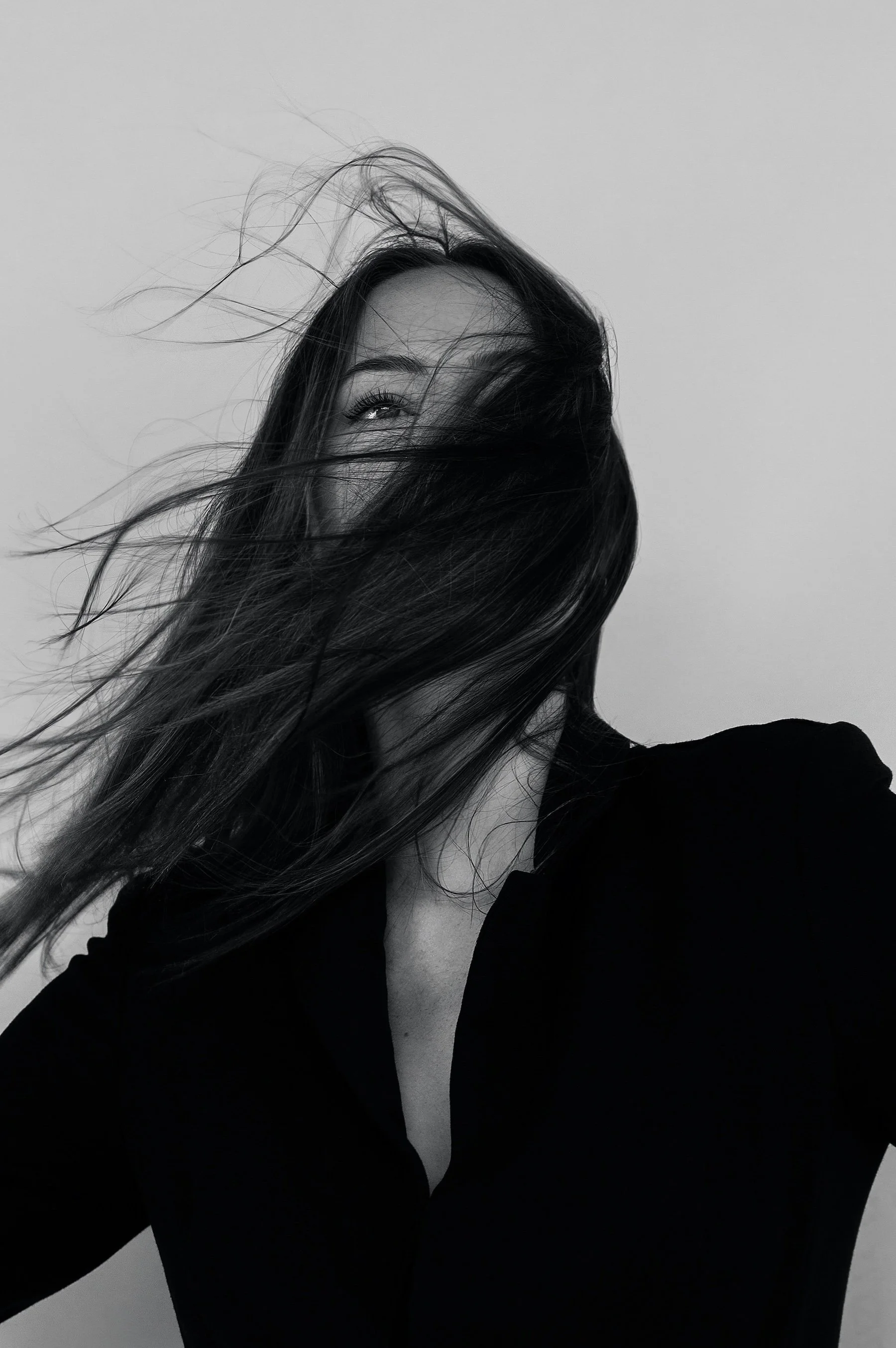 Black and white photo of a woman with long hair partially covering her face, smiling and wearing a dark top, against a plain background.