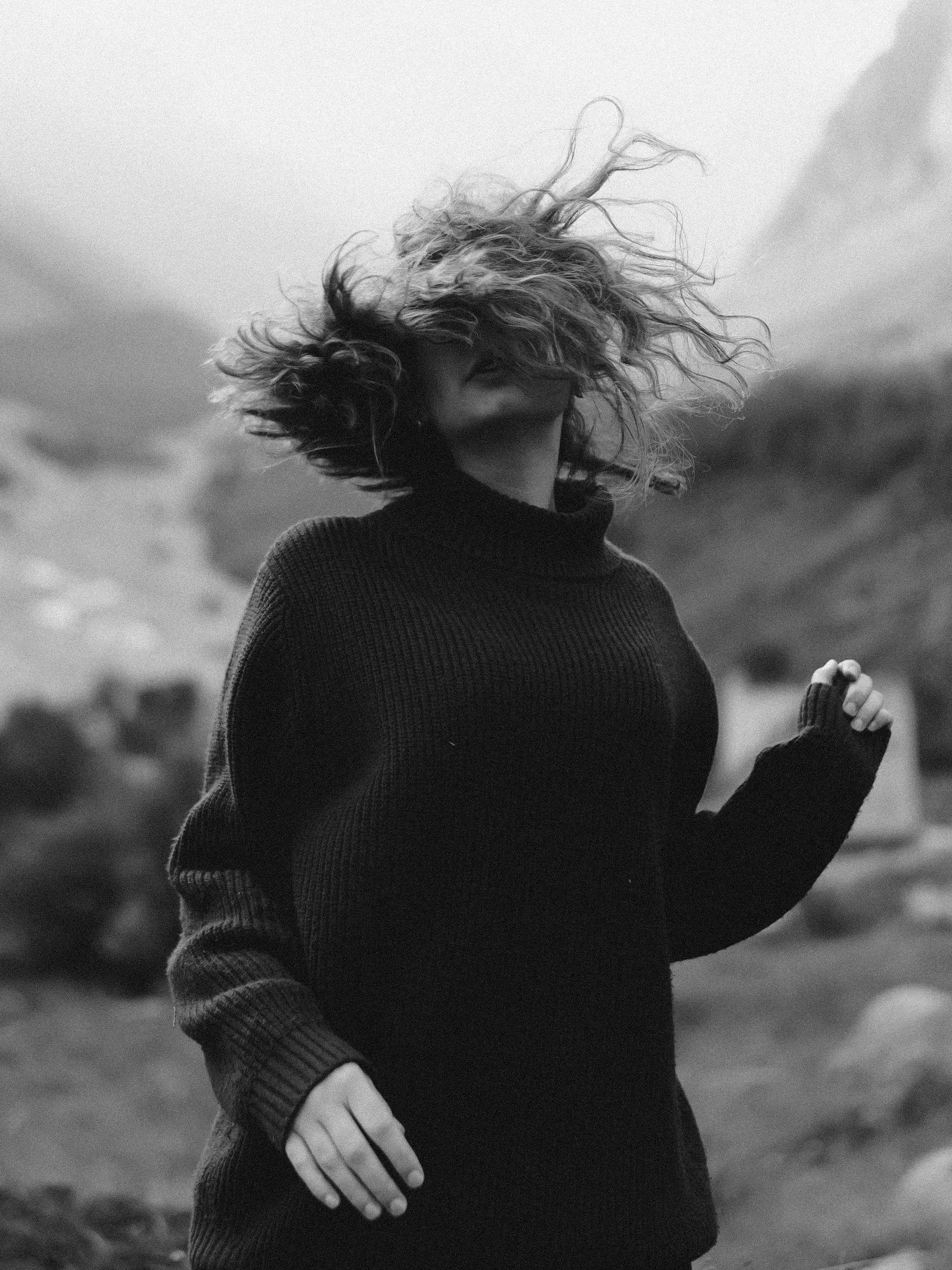 A person with curly hair wearing a dark sweater outdoors with wind blowing hair, blurred natural background.