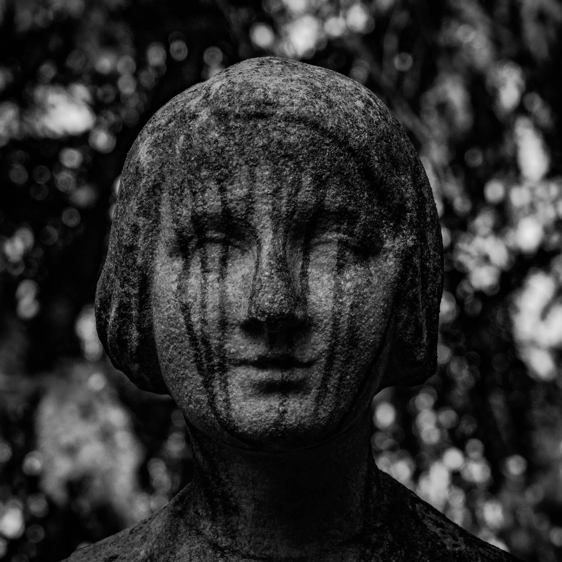 A close-up black and white photograph of a stone statue of a woman's face with closed eyes, with dark streaks resembling tears on its face, and a blurred natural background.