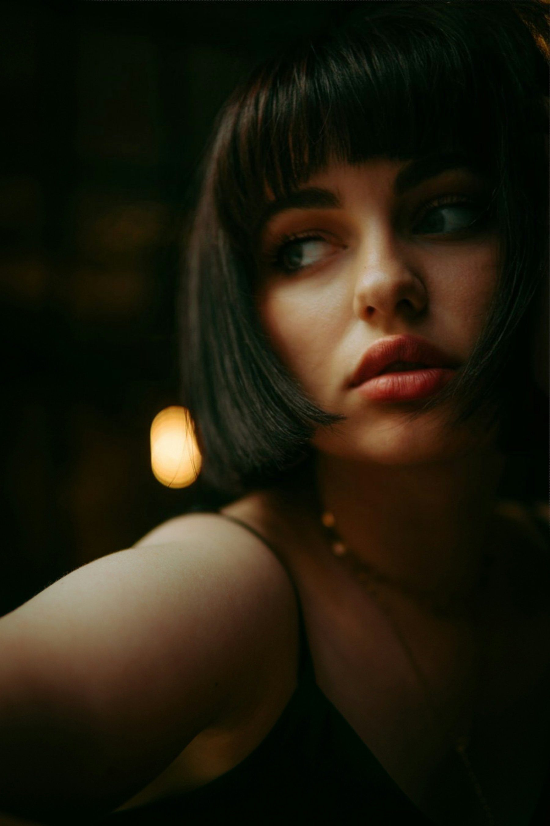 Close-up photo of a woman with dark hair and bangs, looking to the side in a dimly lit setting.