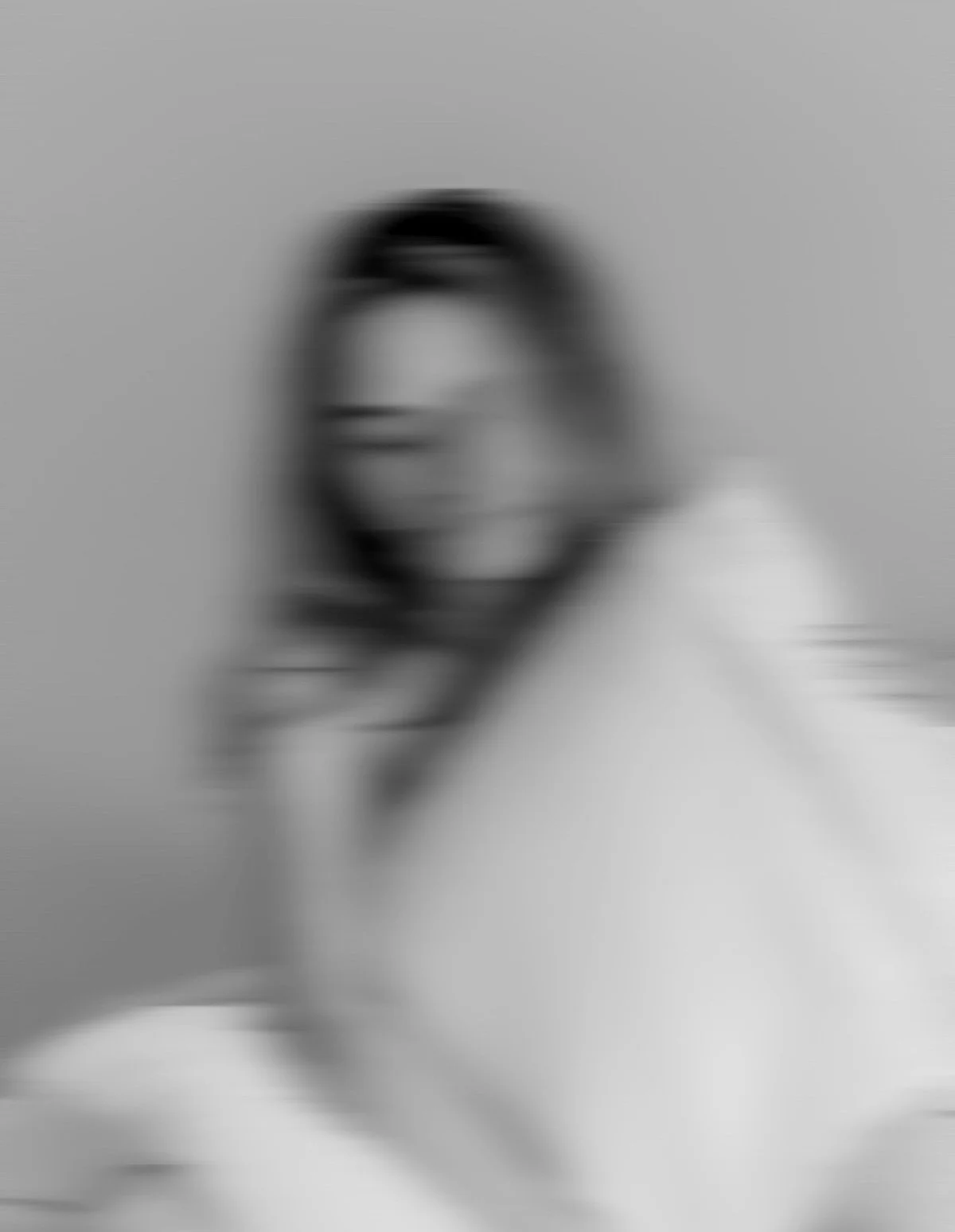 Blurry black and white photo of a woman with elongated features and glasses, smiling with her hair down, against a plain background.