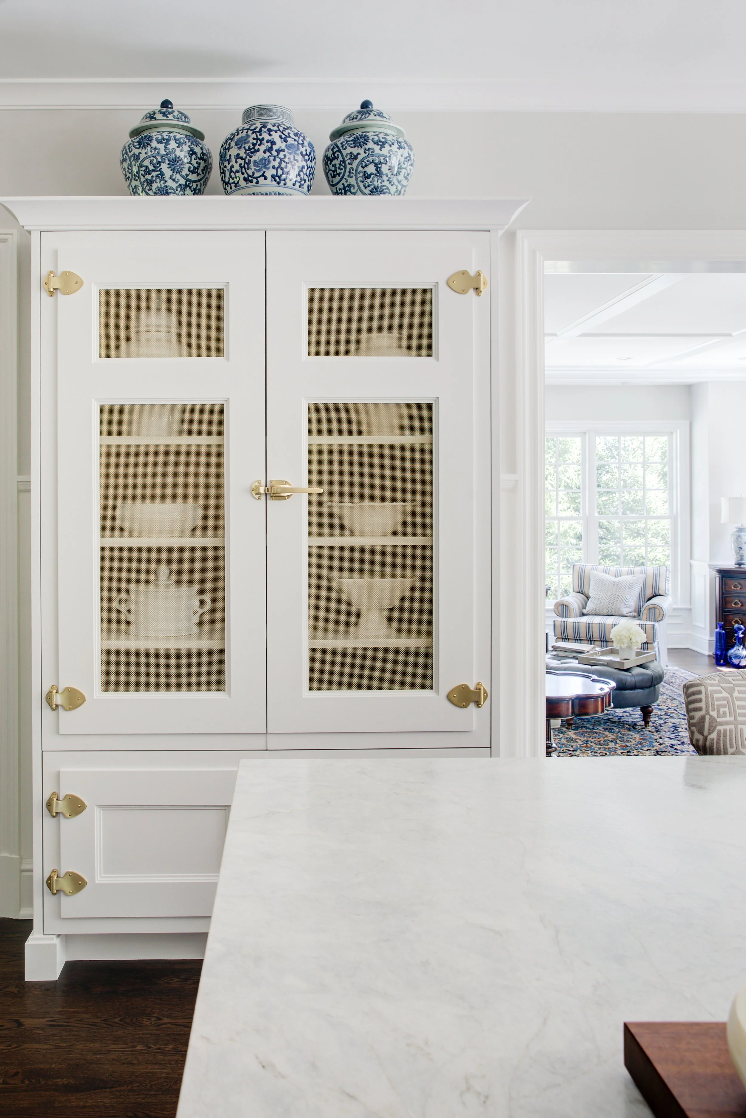 A white kitchen display cabinet with glass-paneled doors and gold hardware, showcasing white and blue ceramic bowls and vases on top, with a living room visible through a doorway in the background.