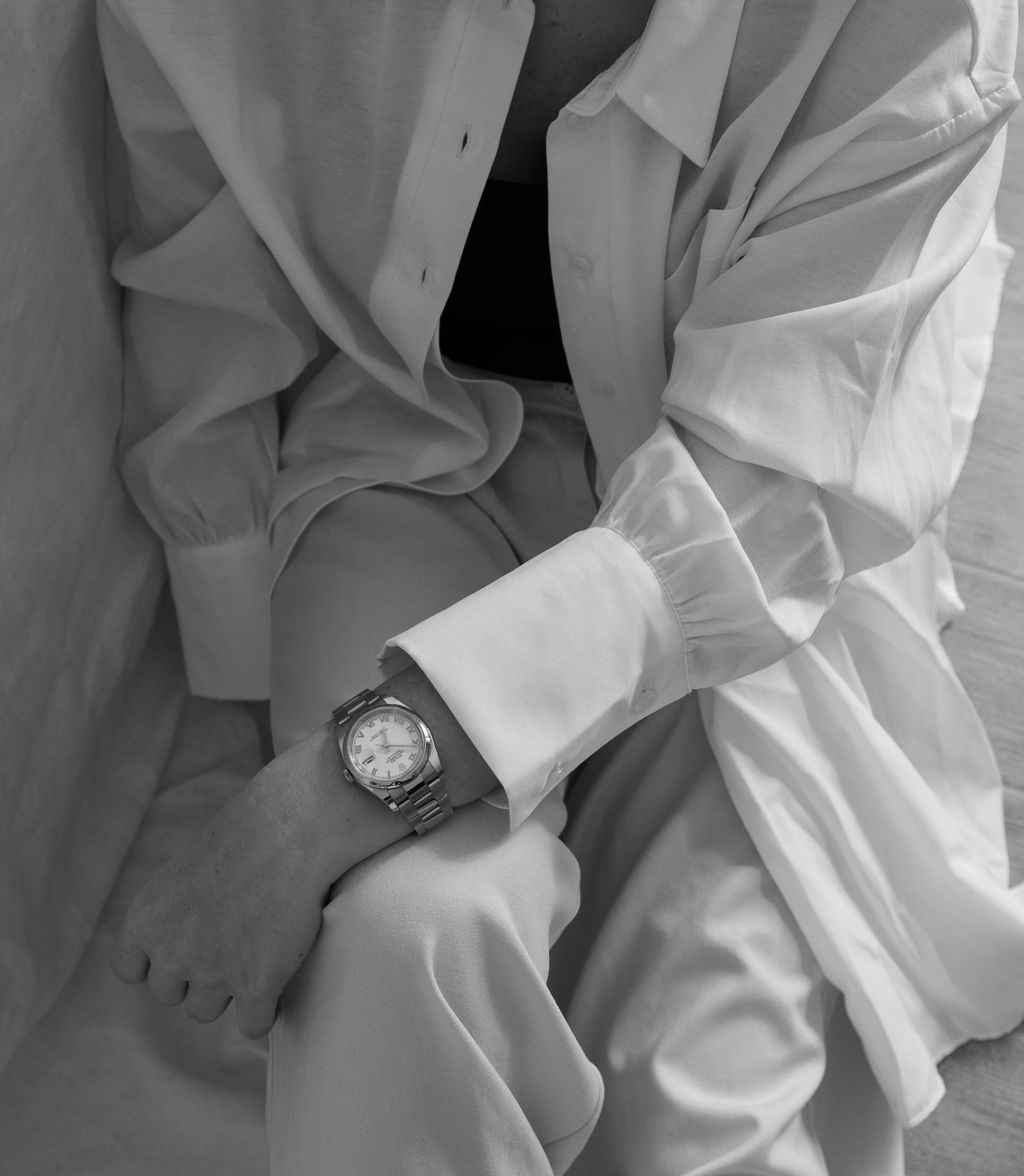 A person wearing a white shirt and light-colored pants, sitting with their arm resting on a surface. They are wearing a watch with a large face on their wrist.