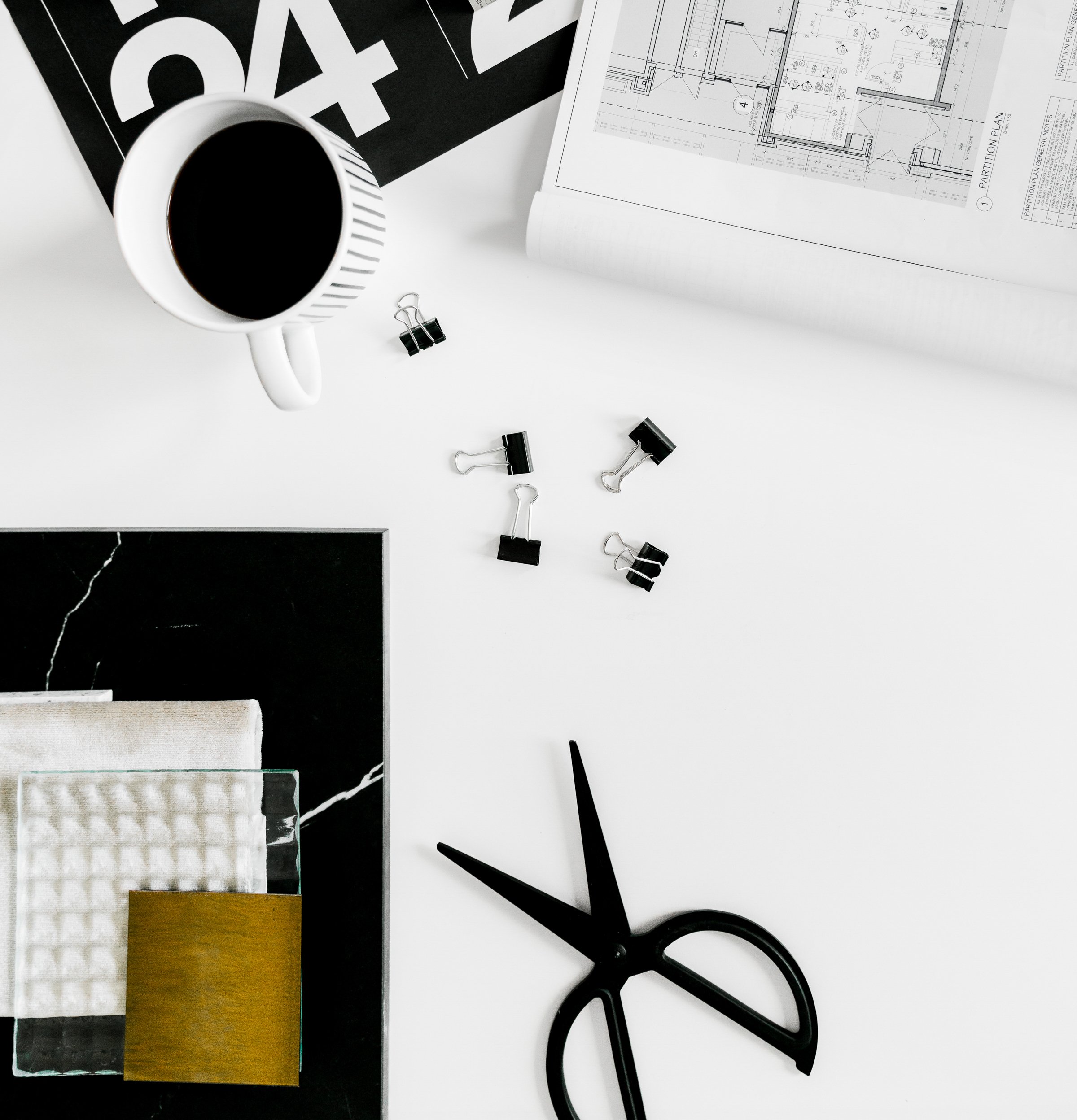 A minimalist black and white work desk with a cup of coffee, architectural blueprints, scattered binder clips, a black pair of scissors, and a black-and-white framed sample board.