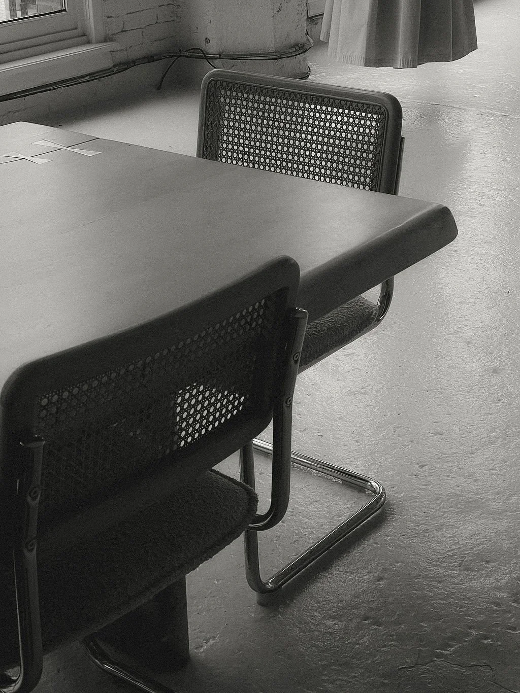 Black mesh-back chairs around a rectangular table in a room with a concrete floor, brick wall, and curtains.