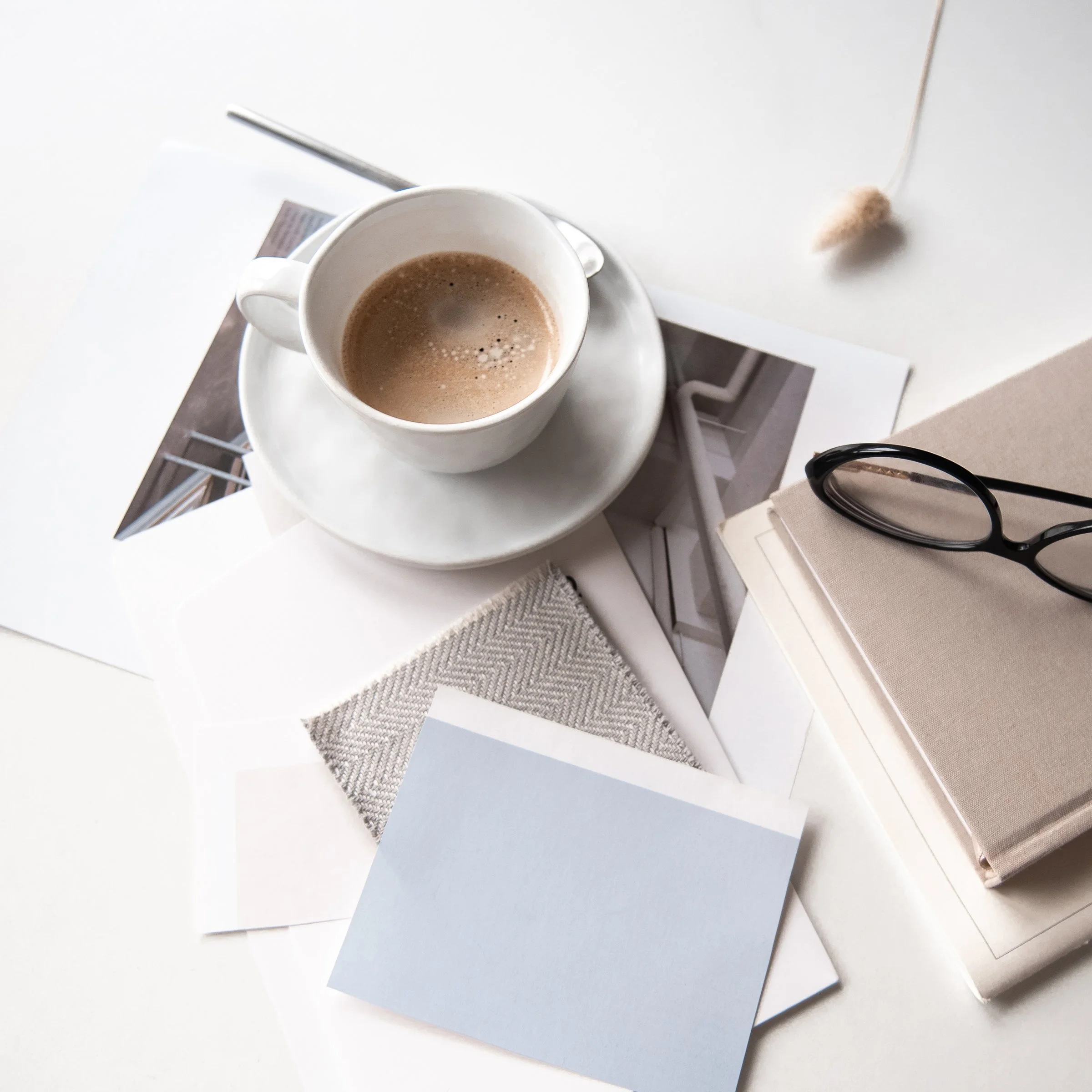 Flat lay of a design workspace with a coffee cup, finish swatches, reading glasses, and a notebook in a neutral palette.