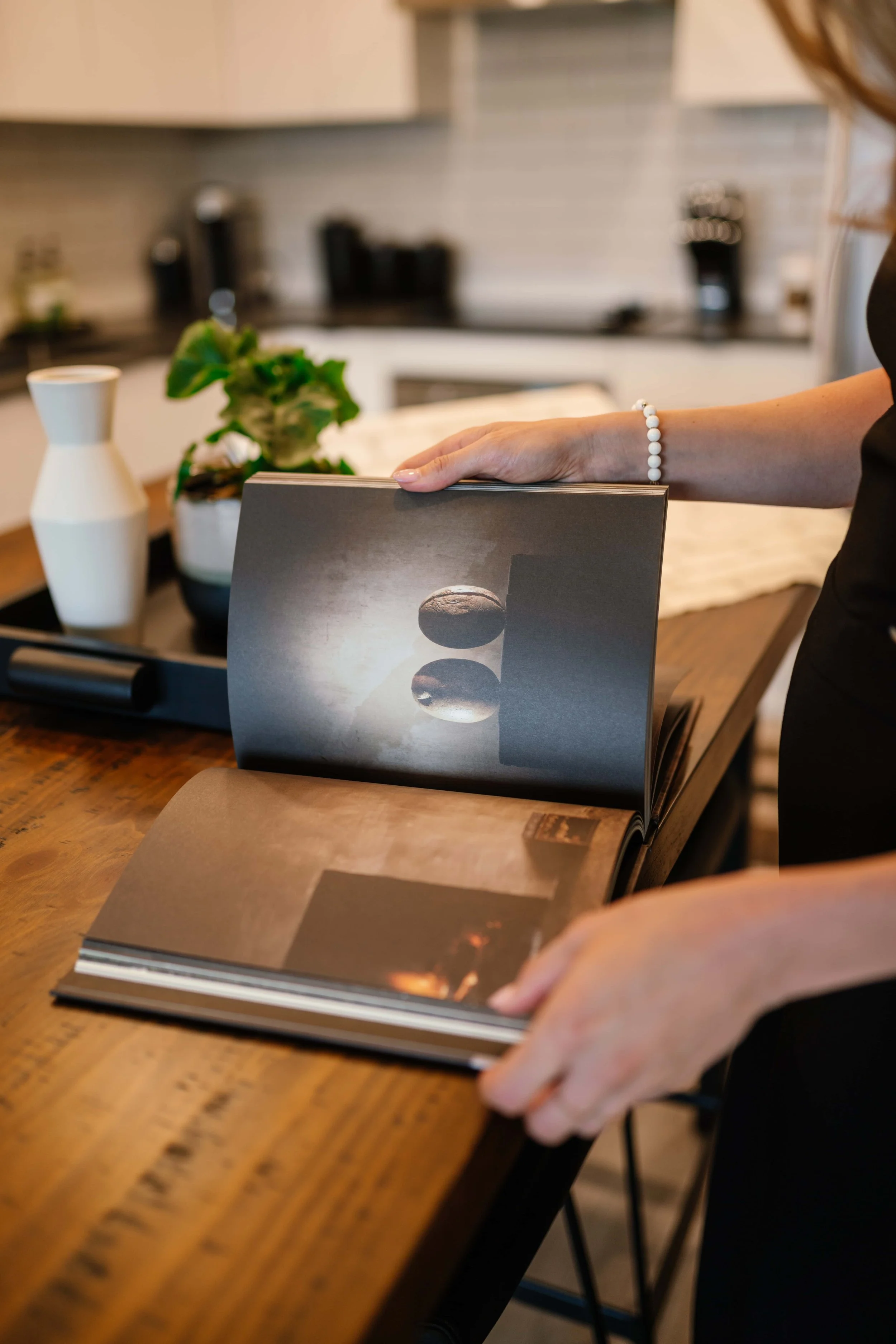 Person flipping through a photo book on a wooden table, showing an image of two spheres and a shadow in a monochrome setting with a light source behind.
