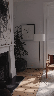 A cozy living room corner with a white floor lamp, a large potted plant, a framed black-and-white artwork, a brown armchair, and hardwood floors with sunlight streaming in.