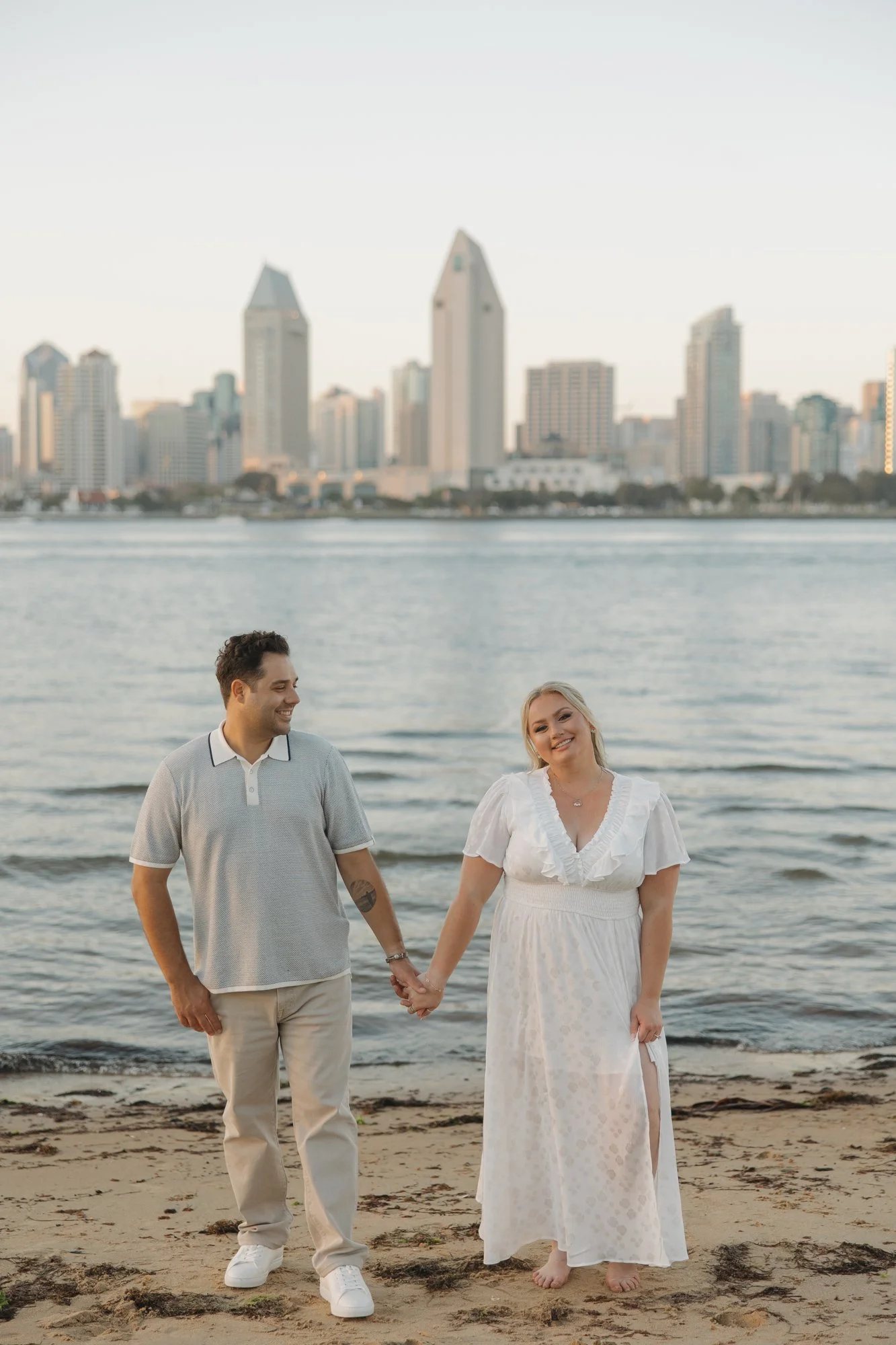 Engaged couple walking hand in hand on the bay beach with the downtown San Diego skyline behind them