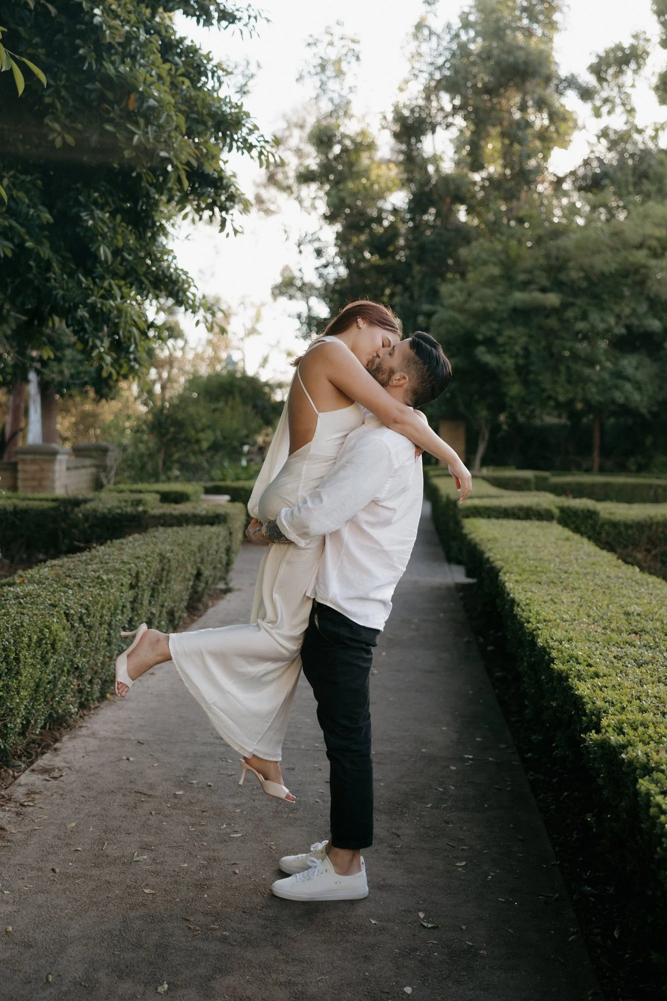 Man lifting his fiancee for a kiss on a hedge-lined garden path in Balboa Park