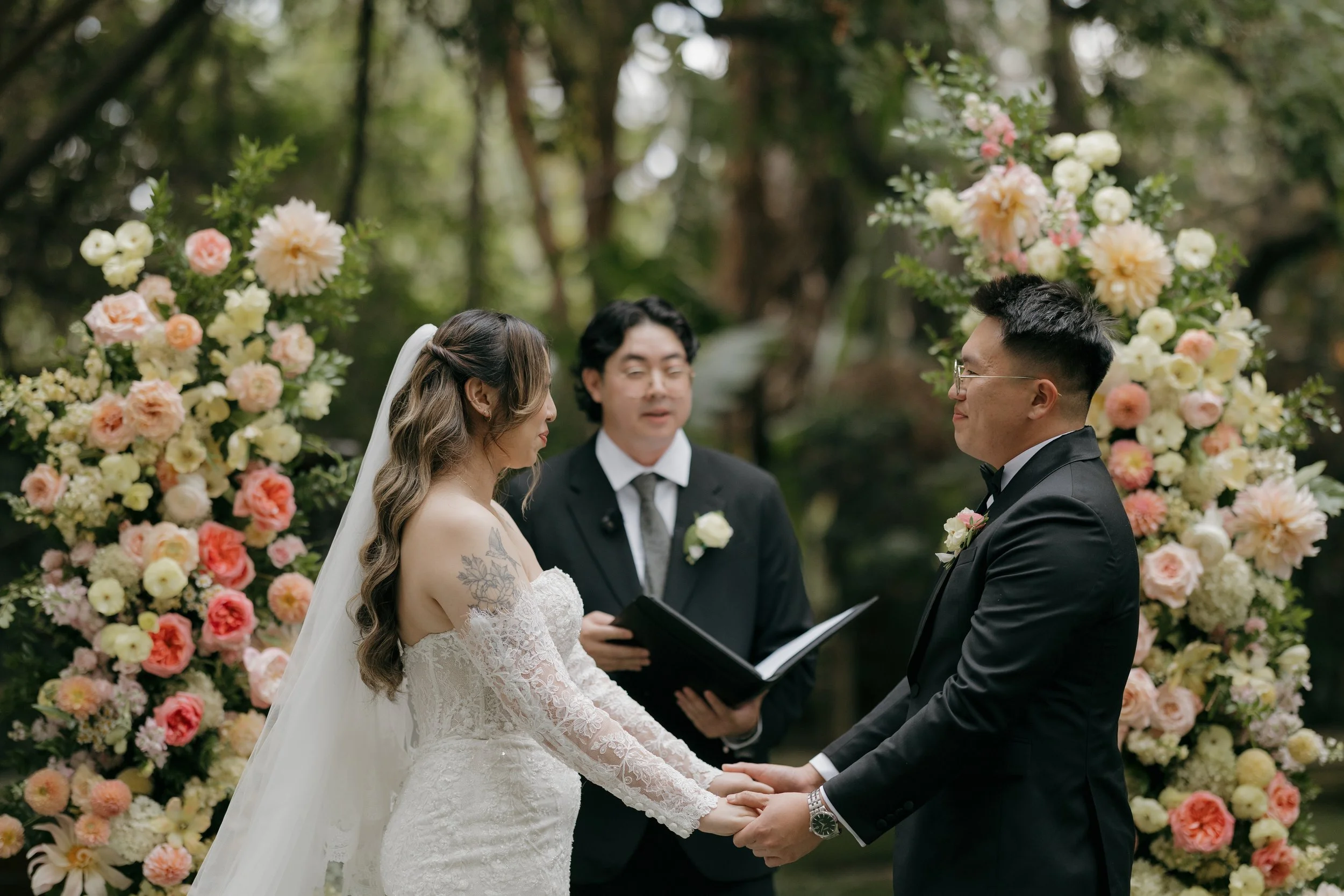 Nancy and Ken holding hands and exchanging vows beneath rose floral arches in San Marcos