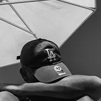 Person lying under a large umbrella, wearing a Los Angeles Dodgers cap, resting with their arm over their eyes.