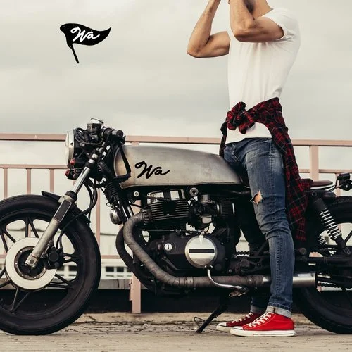 A person standing next to a vintage motorcycle on a bridge, wearing a white t-shirt, ripped jeans, red sneakers, and a red plaid shirt tied around their waist. There is a small black flag with "Wa" on it attached to the motorcycle.
