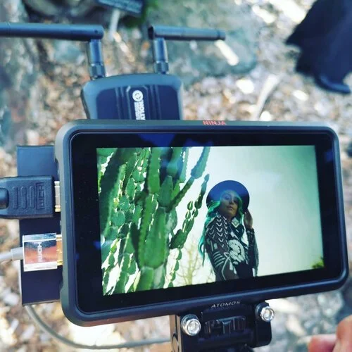 A camera recording a woman standing outdoors near a cactus plant, with trees and ground visible in the background.