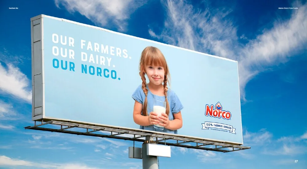 Billboard advertisement for Norco dairy featuring a young girl with red hair in pigtails holding a glass of milk against a blue sky background. The text on the billboard reads 'Our farmers. Our dairy. Our Norco.' and emphasizes farmer ownership.