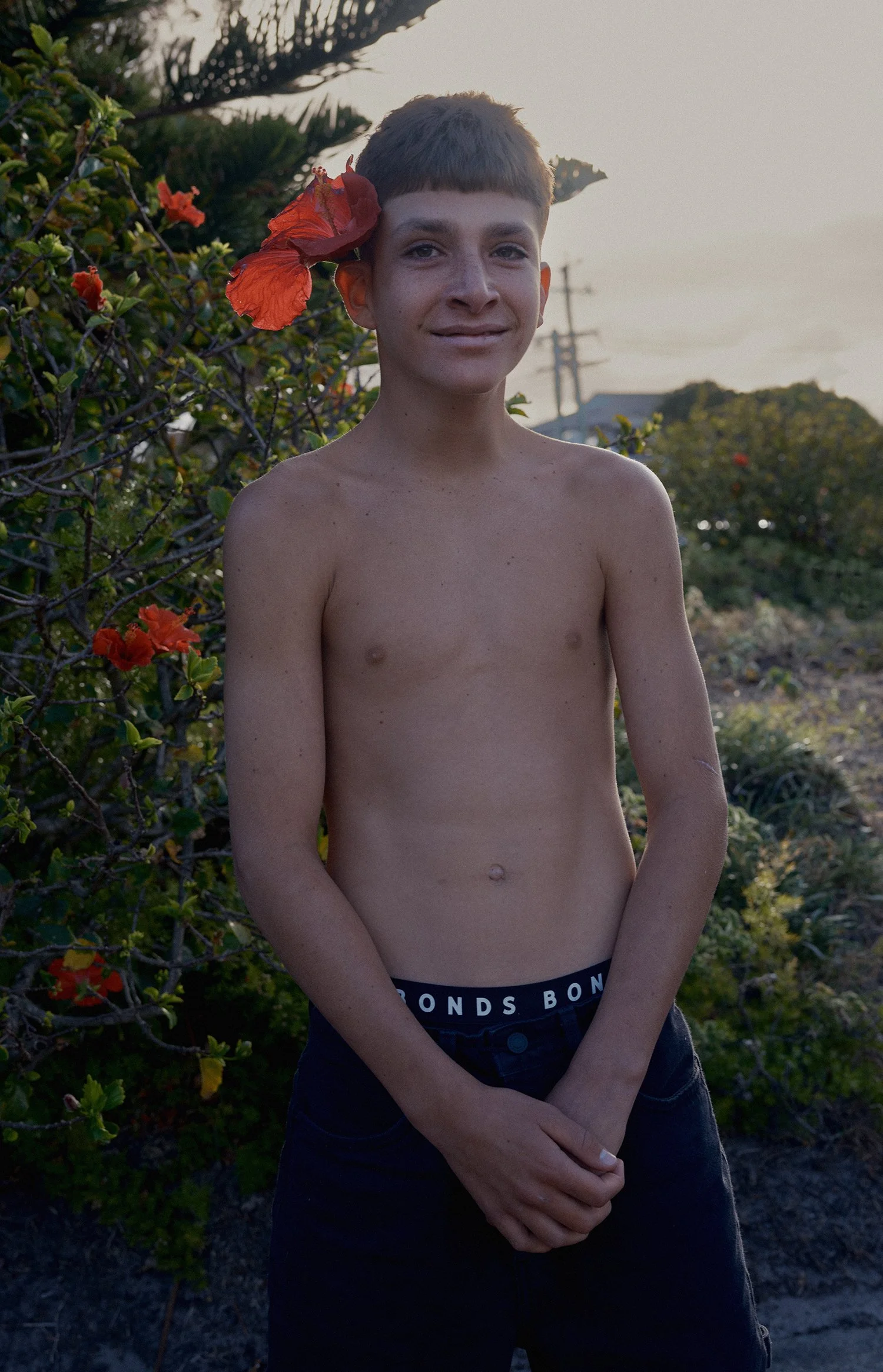 Shirtless person with a red flower behind their ear, standing outdoors with bushes in the background.