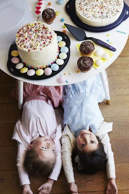 Two children lying under a table with cakes and cupcakes on top, decorated with sprinkles and colorful candy.