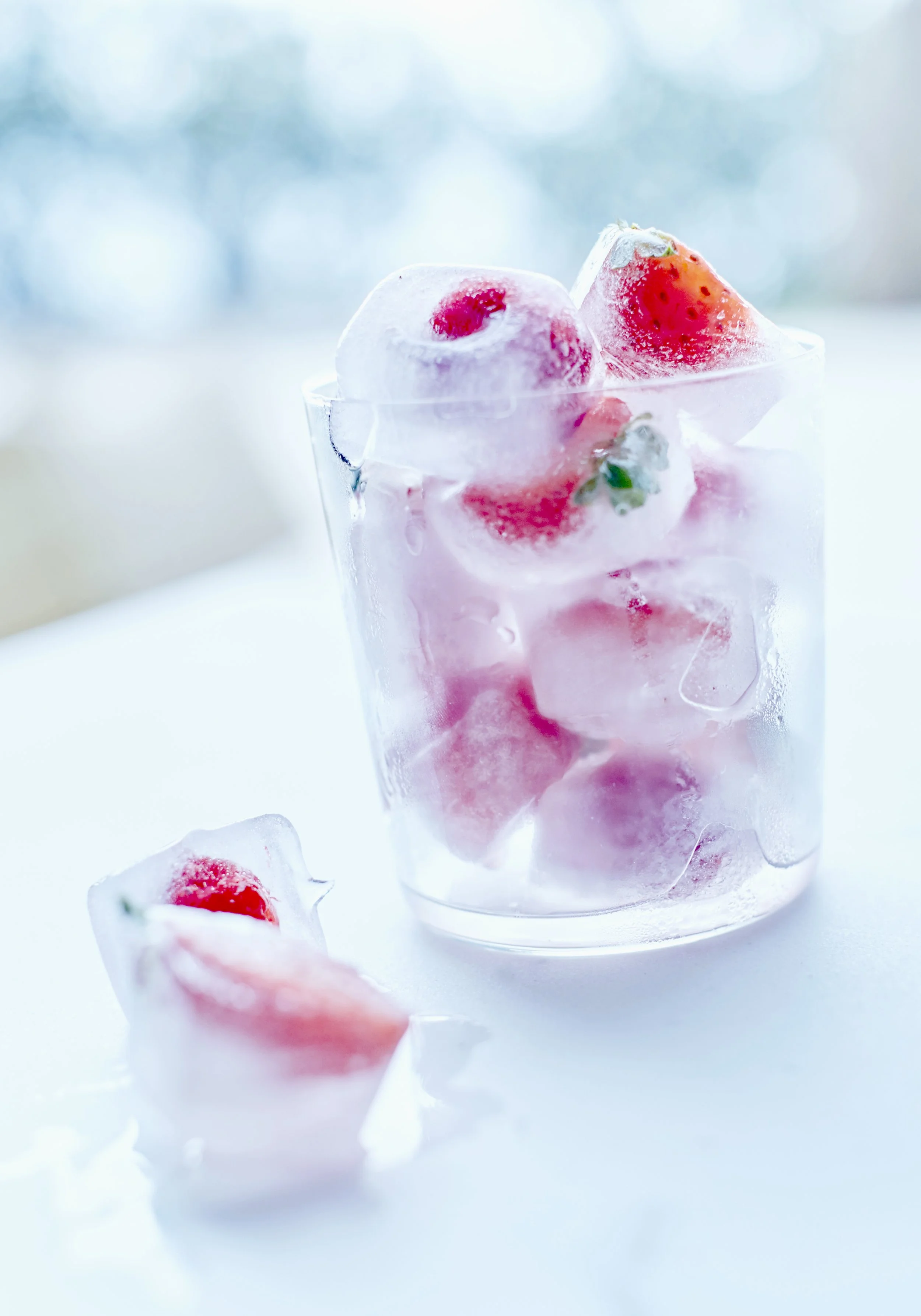 Glass with ice cubes containing strawberries on a white surface.