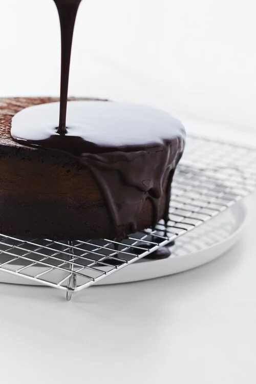 Chocolate ganache being poured over a cake on a wire cooling rack.