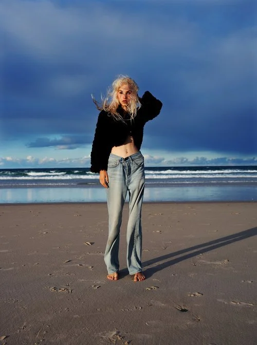 Person standing on a beach, wearing a black sweater and jeans, with the ocean and a partly cloudy sky in the background.