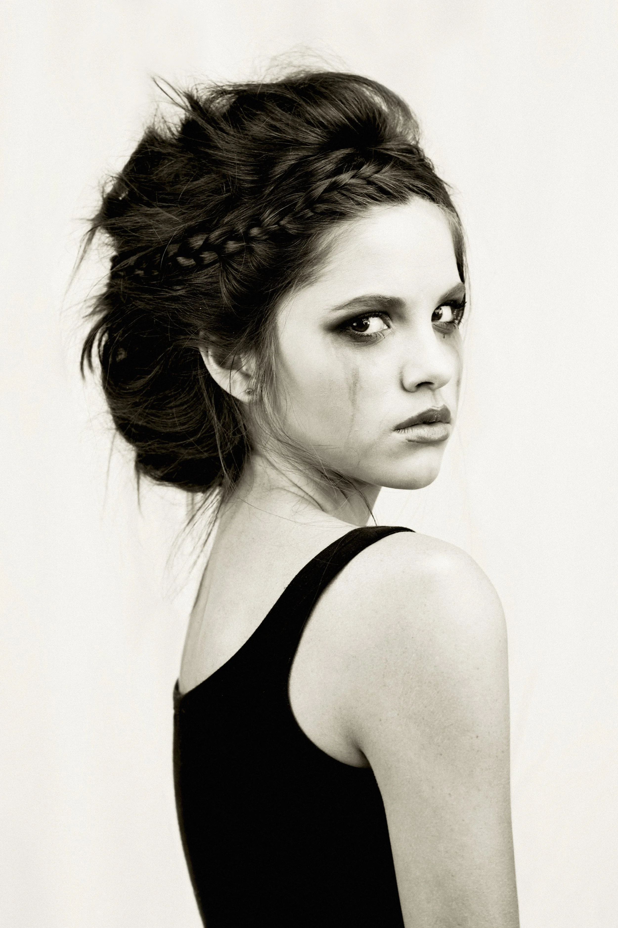 Black and white portrait of a woman wearing a black dress, with braided hair and intense expression, looking over her shoulder.