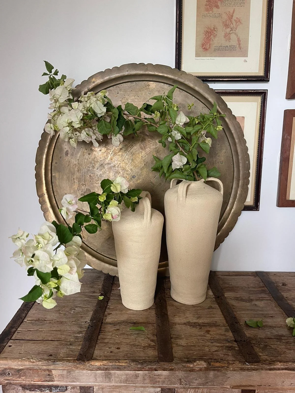 Two tall beige vases with leaves and white flowers, placed in front of a large silver tray and framed pictures on a wall.